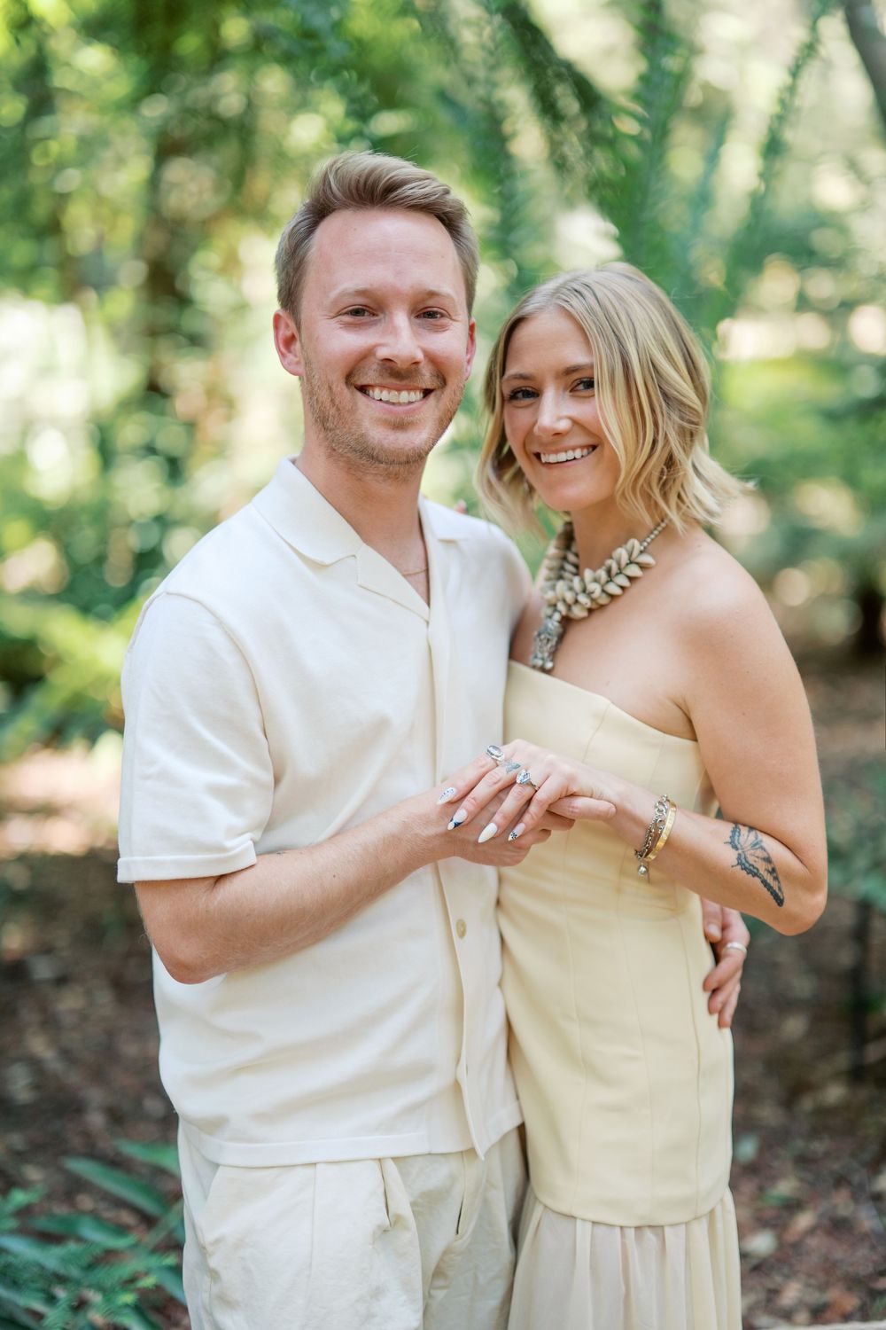 A newly engaged couple poses for a portrait in a lush garden setting wearing coordinated cream-colored outfits.