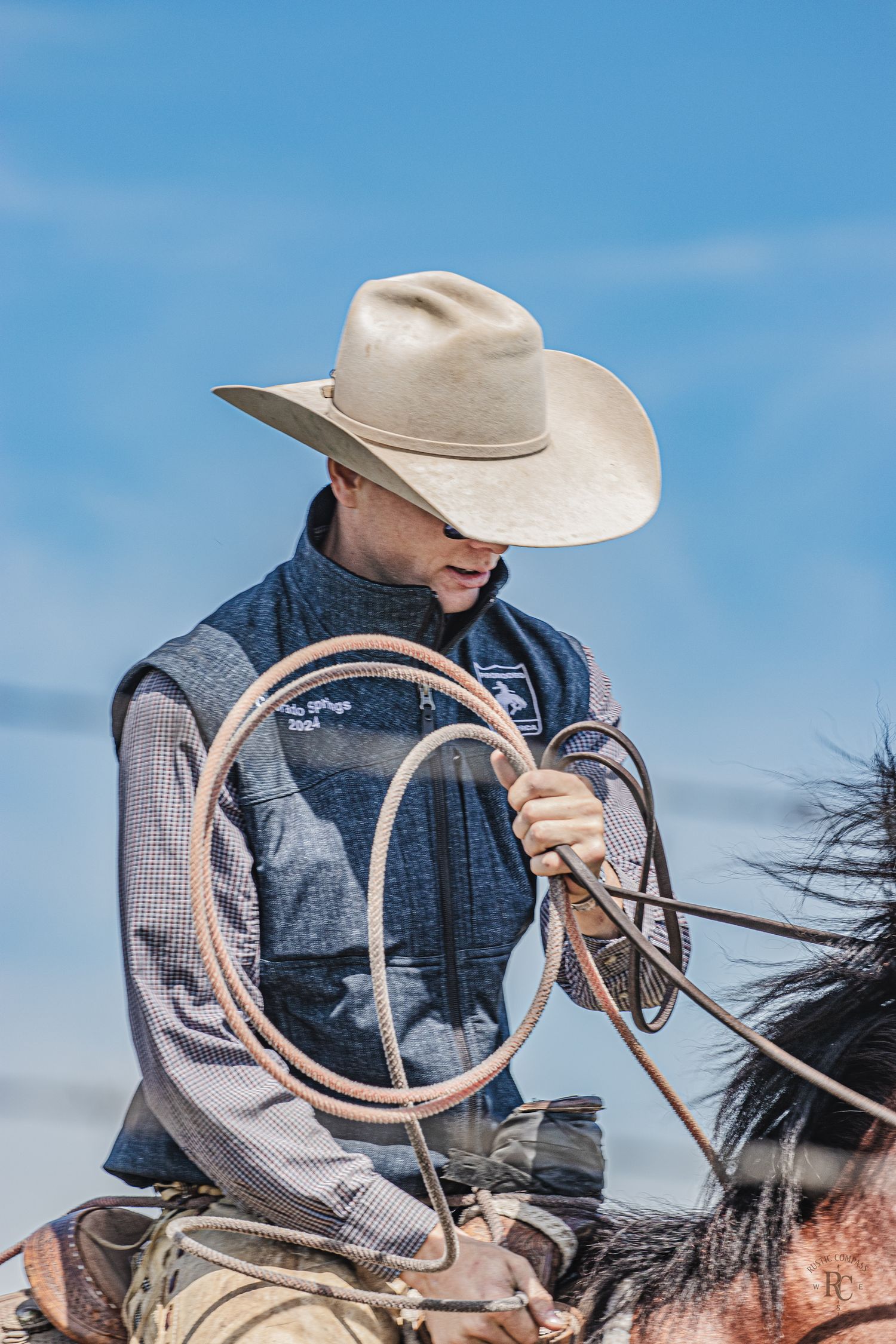 Western style rider wearing cowboy hat and holding lasso on horseback against blue sky background.