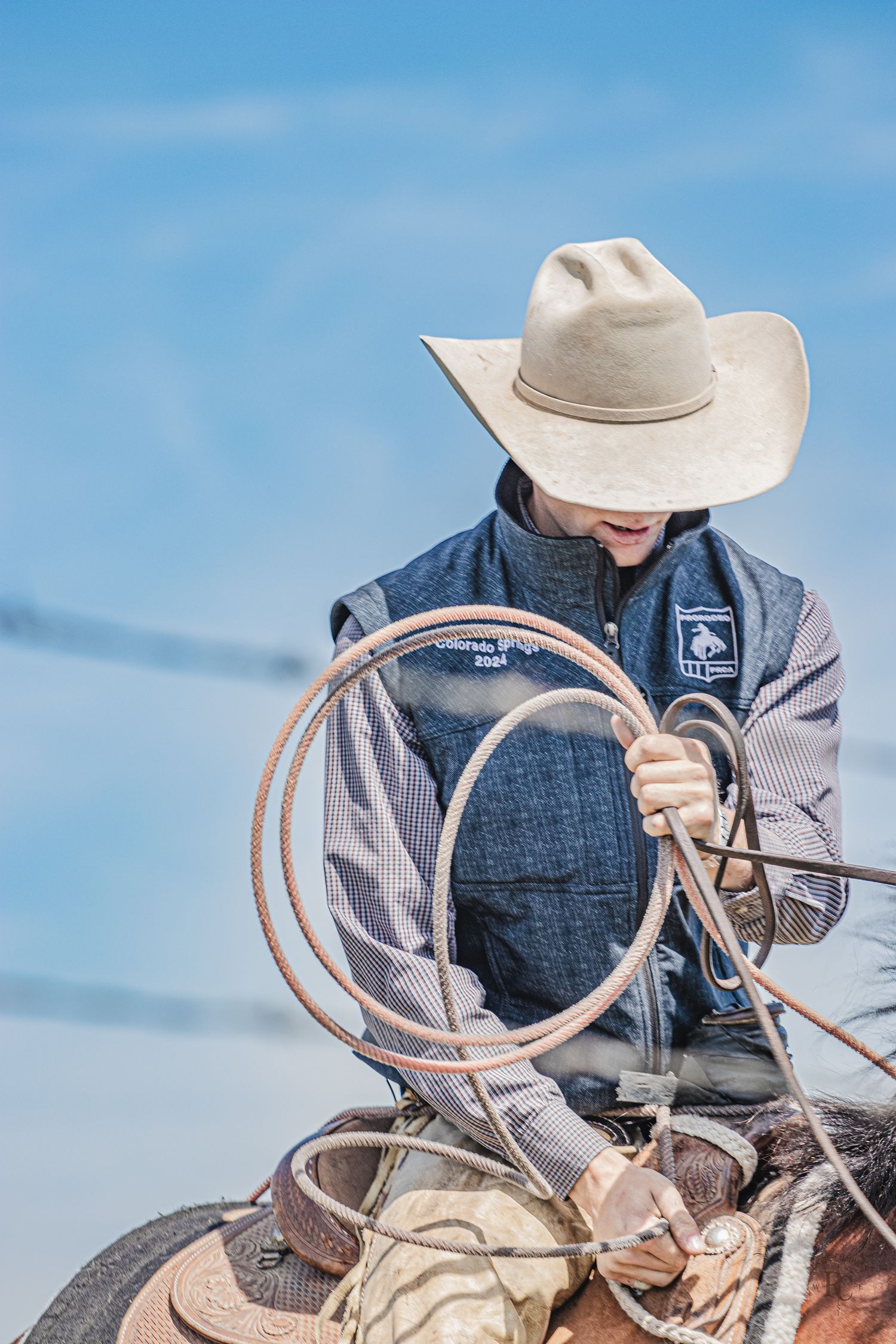 Western cowboy on horseback practices lassoing technique with golden rope against a bright blue sky background.