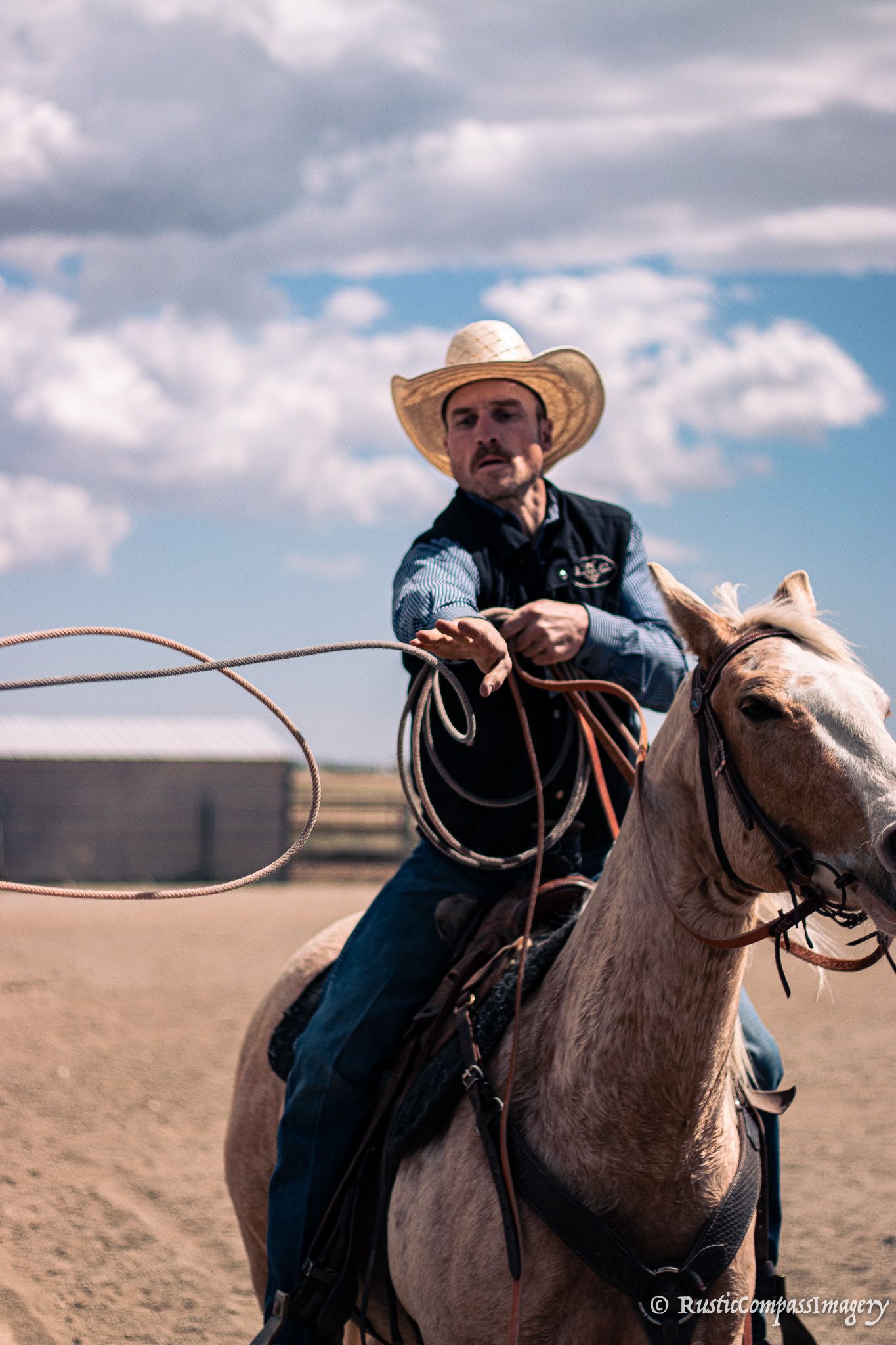 A cowboy wearing a hat holds a lasso while riding a light-colored horse against a cloudy blue sky.