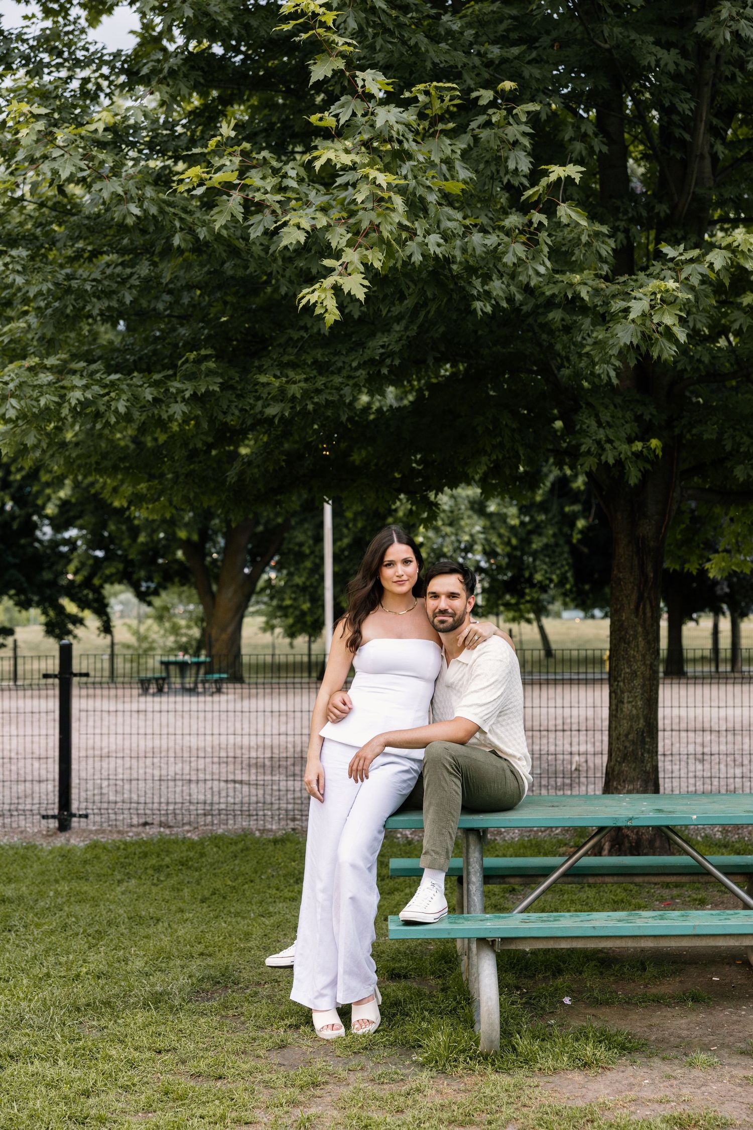 A couple sits together on a green picnic bench in a serene park setting surrounded by leafy trees.