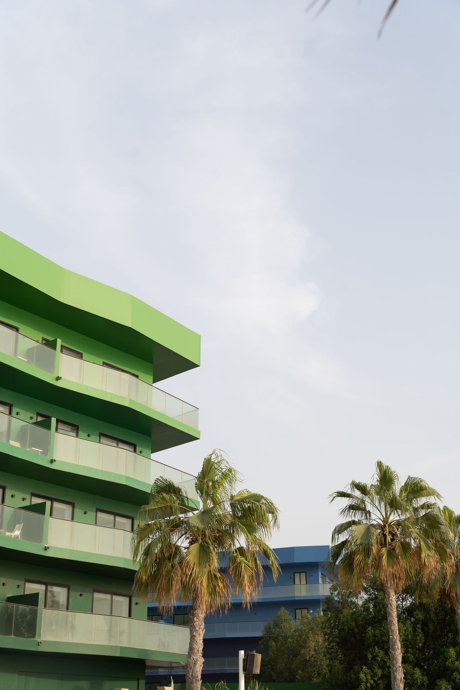 A green modern apartment building with balconies stands next to palm trees against a cloudy sky.