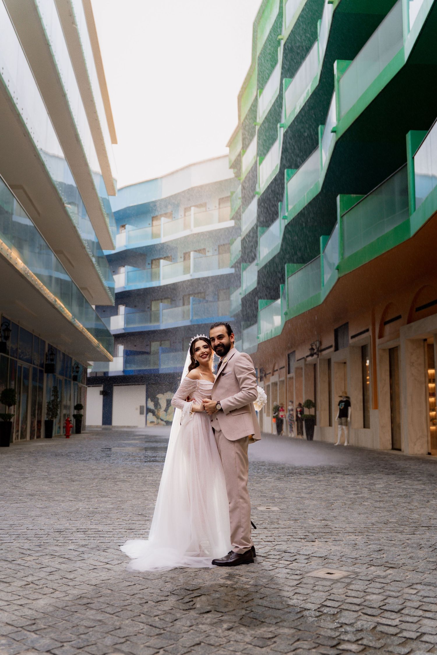 A couple in wedding attire embraces in a modern courtyard with green and white architectural details and cobblestone ground.