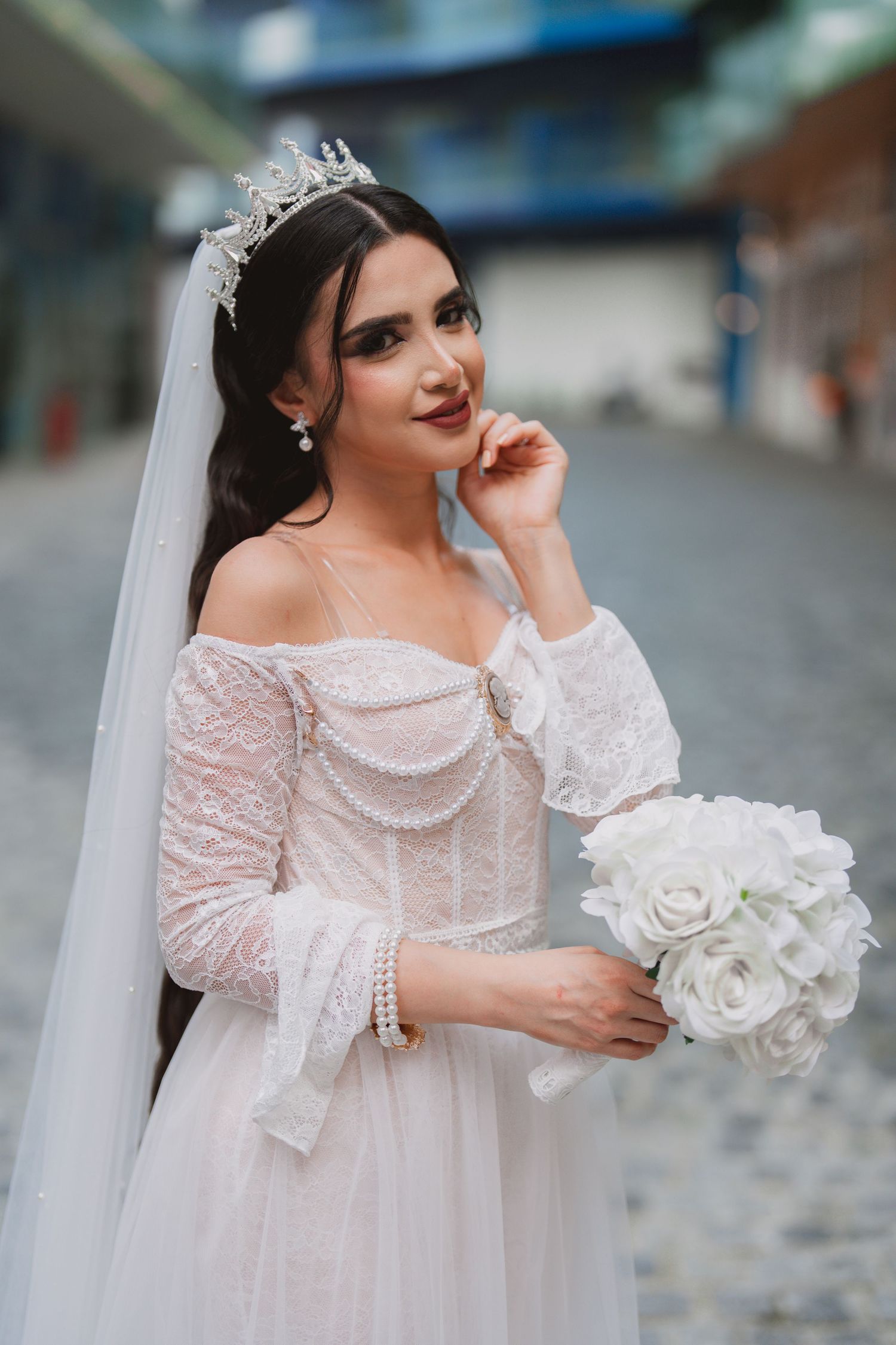 Bride holding white rose bouquet poses in delicate lace wedding dress with flowing veil.