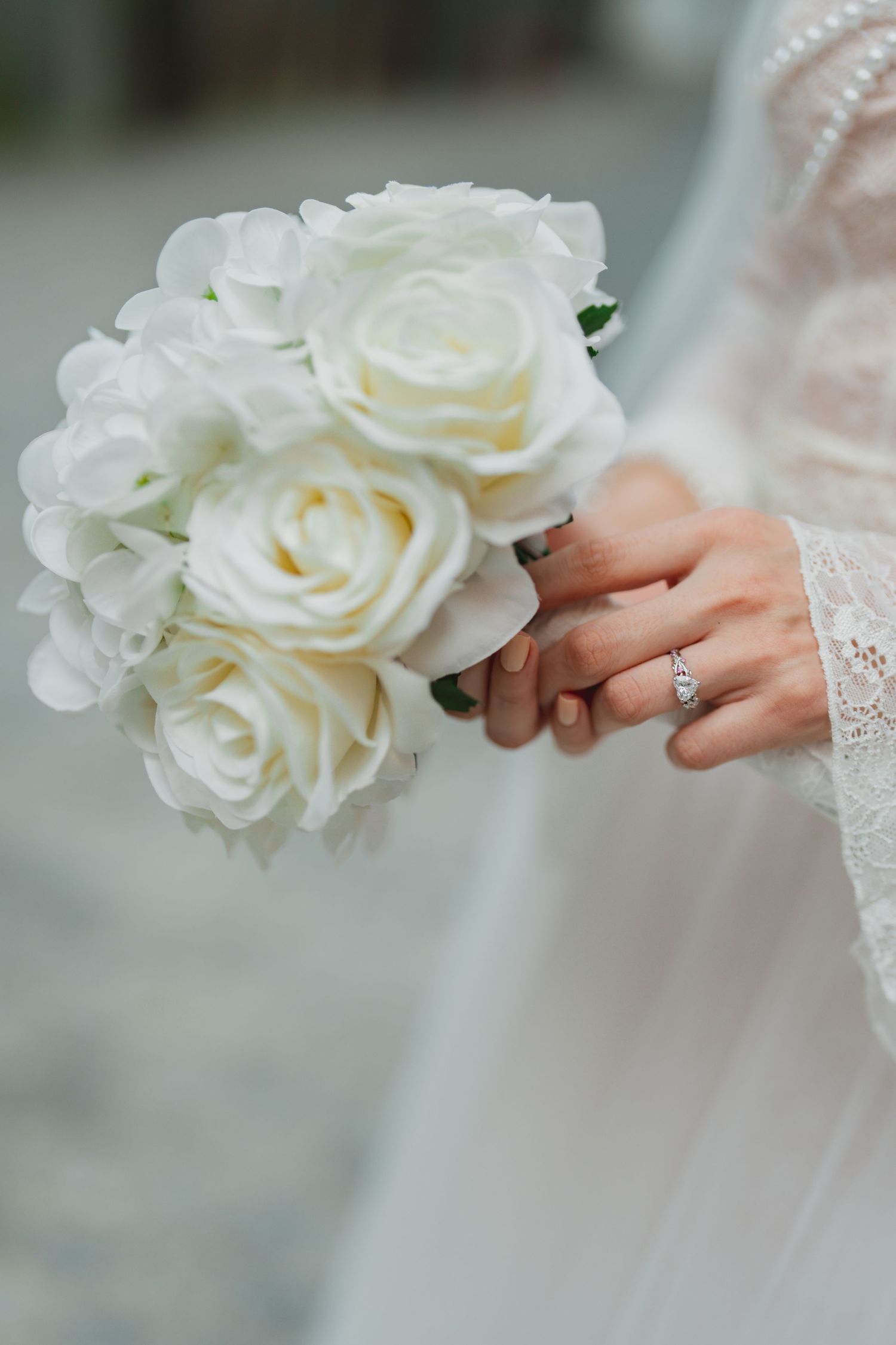 A delicate diamond engagement ring sparkles next to a bouquet of white roses held in soft focus.
