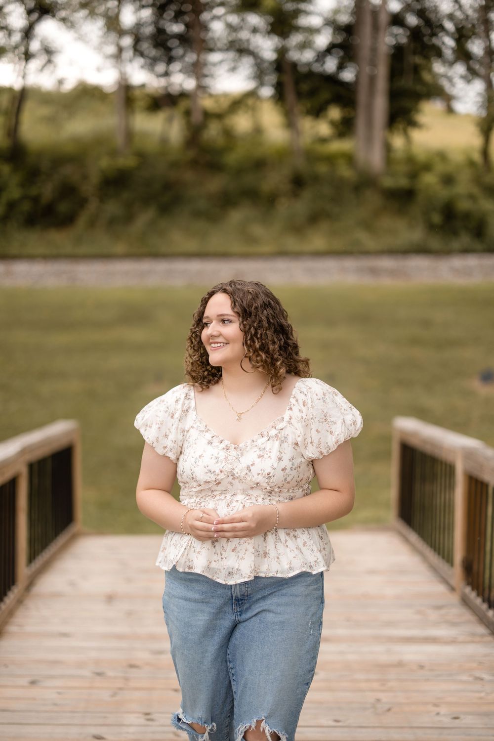 A person stands on a wooden boardwalk wearing a white floral blouse and distressed jeans on a sunny afternoon.