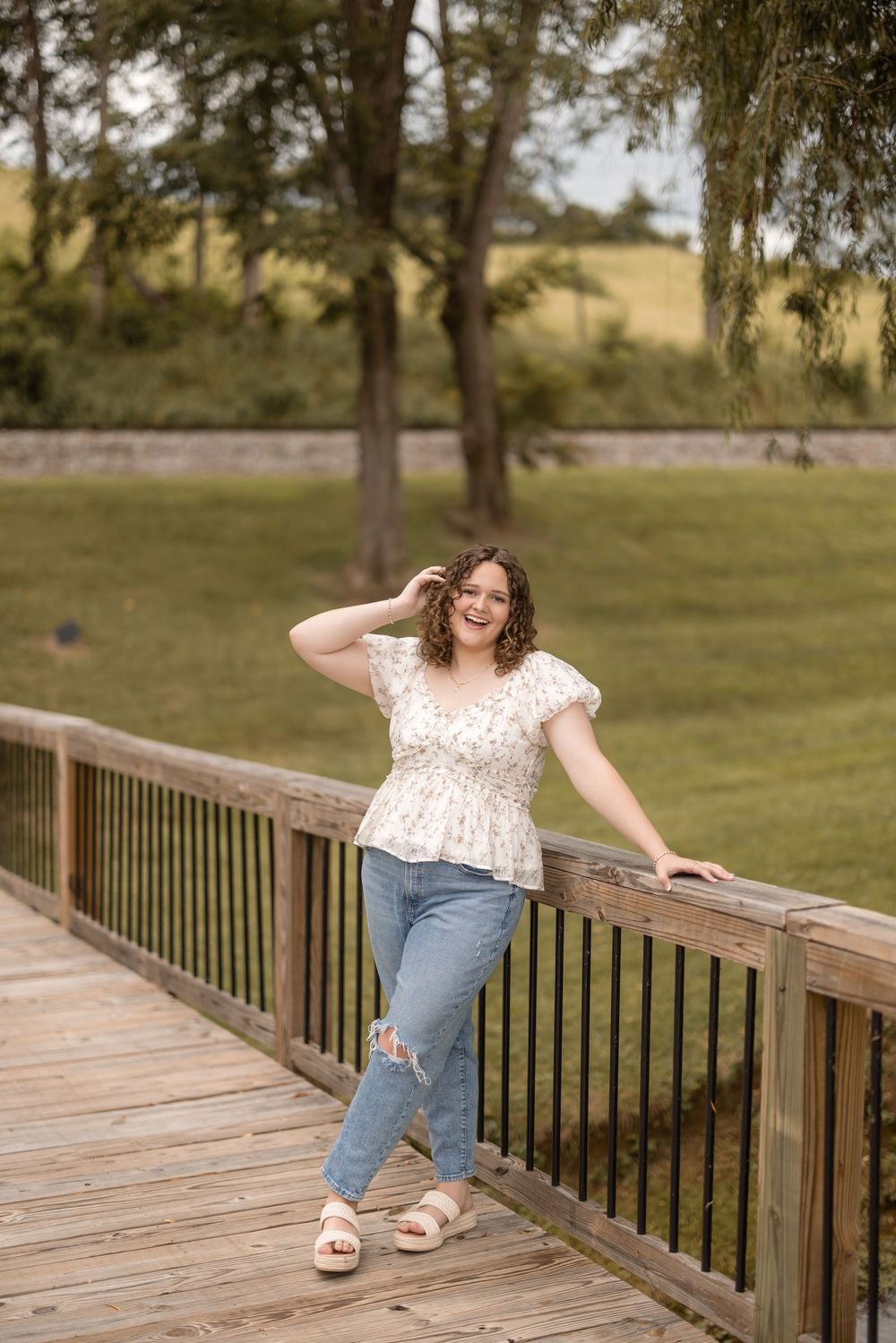 Person in white floral top and jeans poses playfully along a wooden deck railing in a park setting.