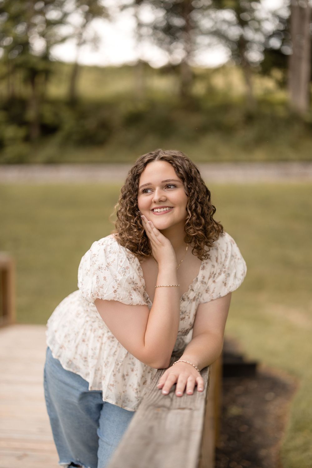 Person in white lace top and jeans sits casually outdoors with scenic natural background during photo session.