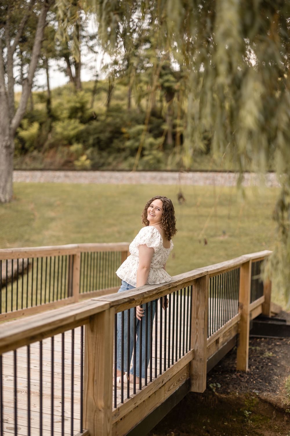 A person in a white top and jeans leans against the railing of a wooden footbridge surrounded by trees.