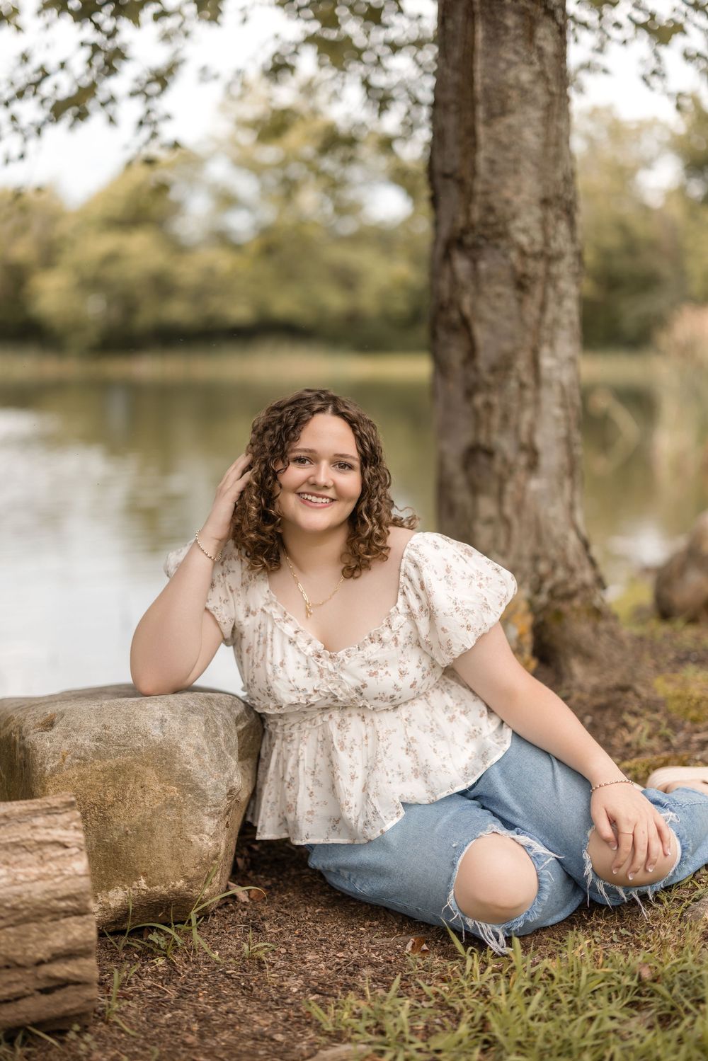 A person sits by a lake wearing a white lace top and ripped jeans while posing next to a tree.
