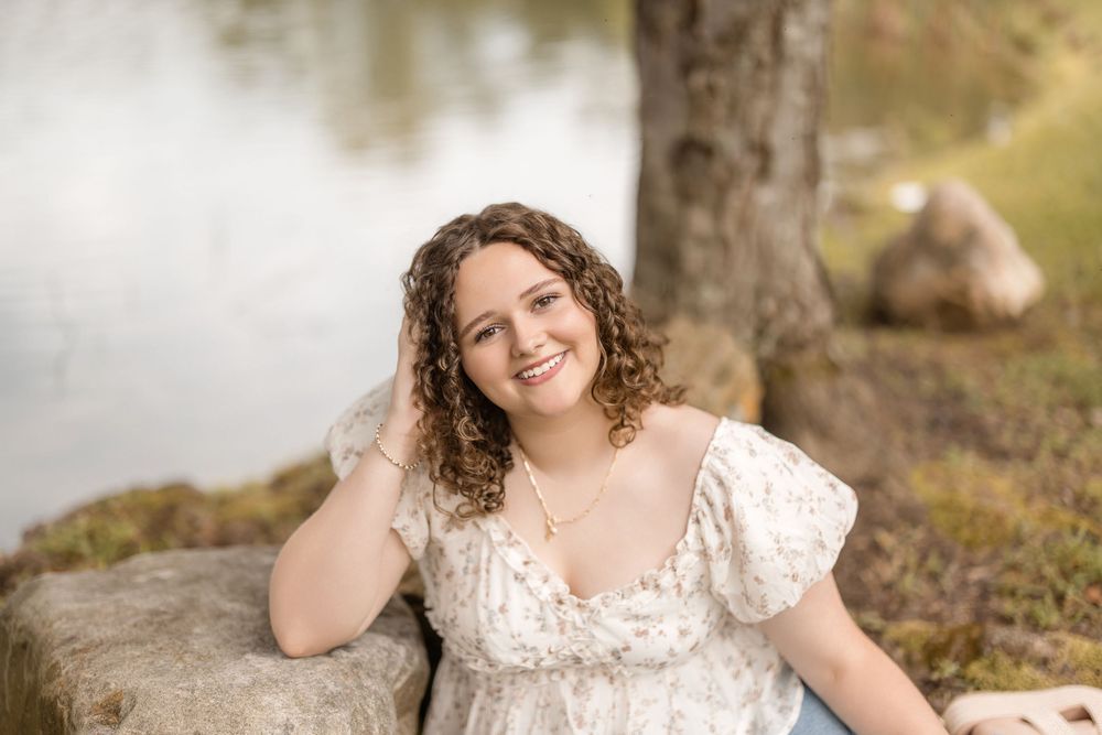 Someone in a white lacy dress sits on a rock by a lake, with curly hair and a warm natural smile.