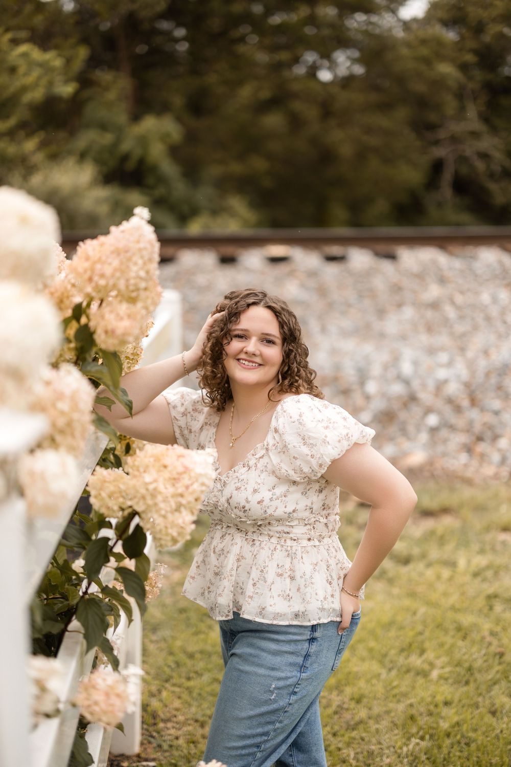 A person in a floral peplum top and jeans stands in a garden with blooming hydrangeas against a stone wall.