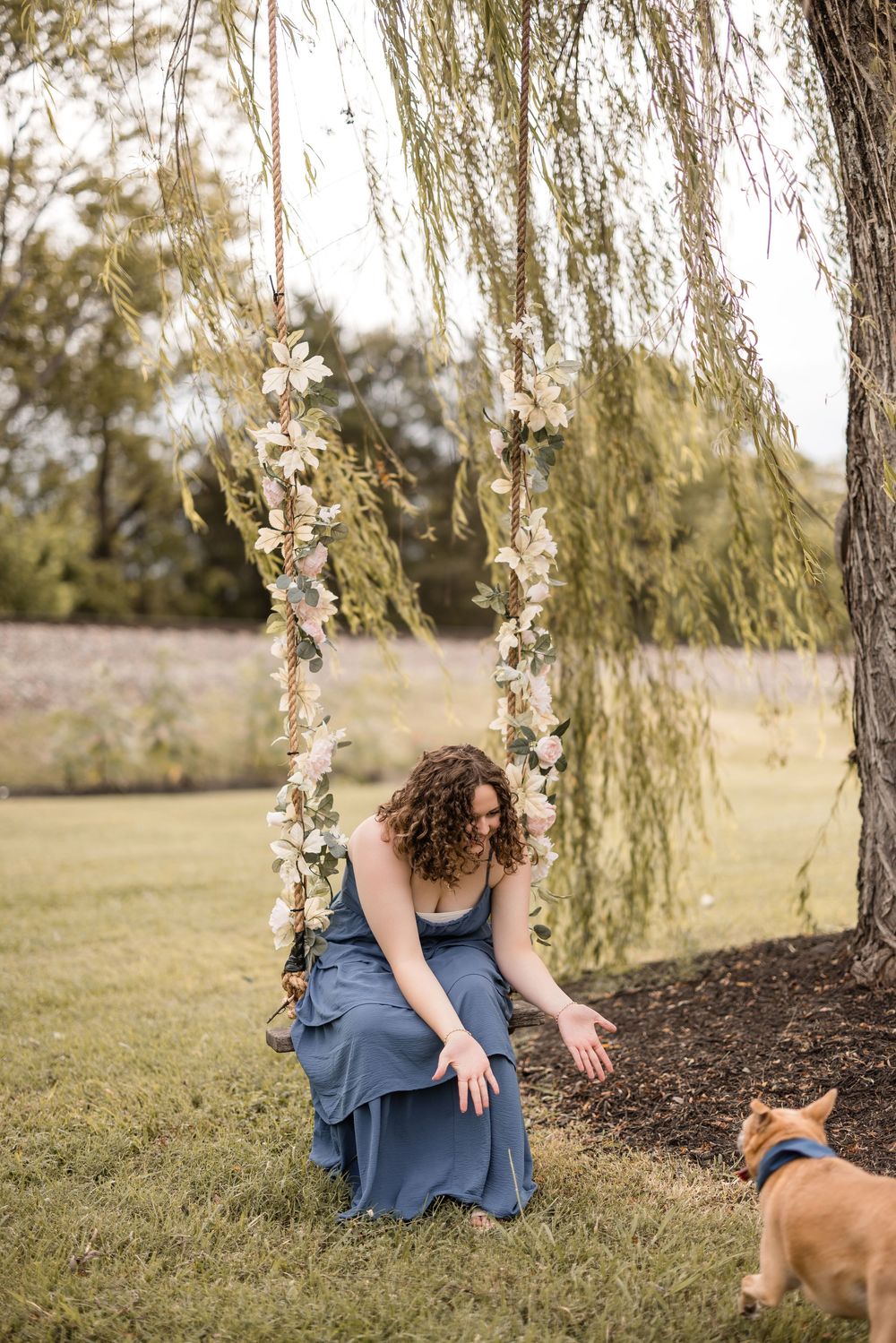 A person in a flowing blue dress kneels beneath a willow tree while a dog watches nearby on a summer afternoon.