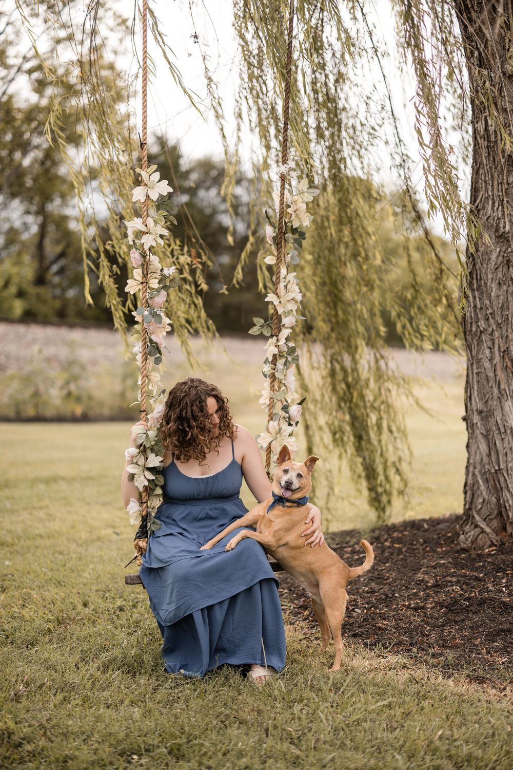 A tranquil outdoor scene featuring someone in a blue dress sitting on a decorated swing with a tan dog under a willow tree.