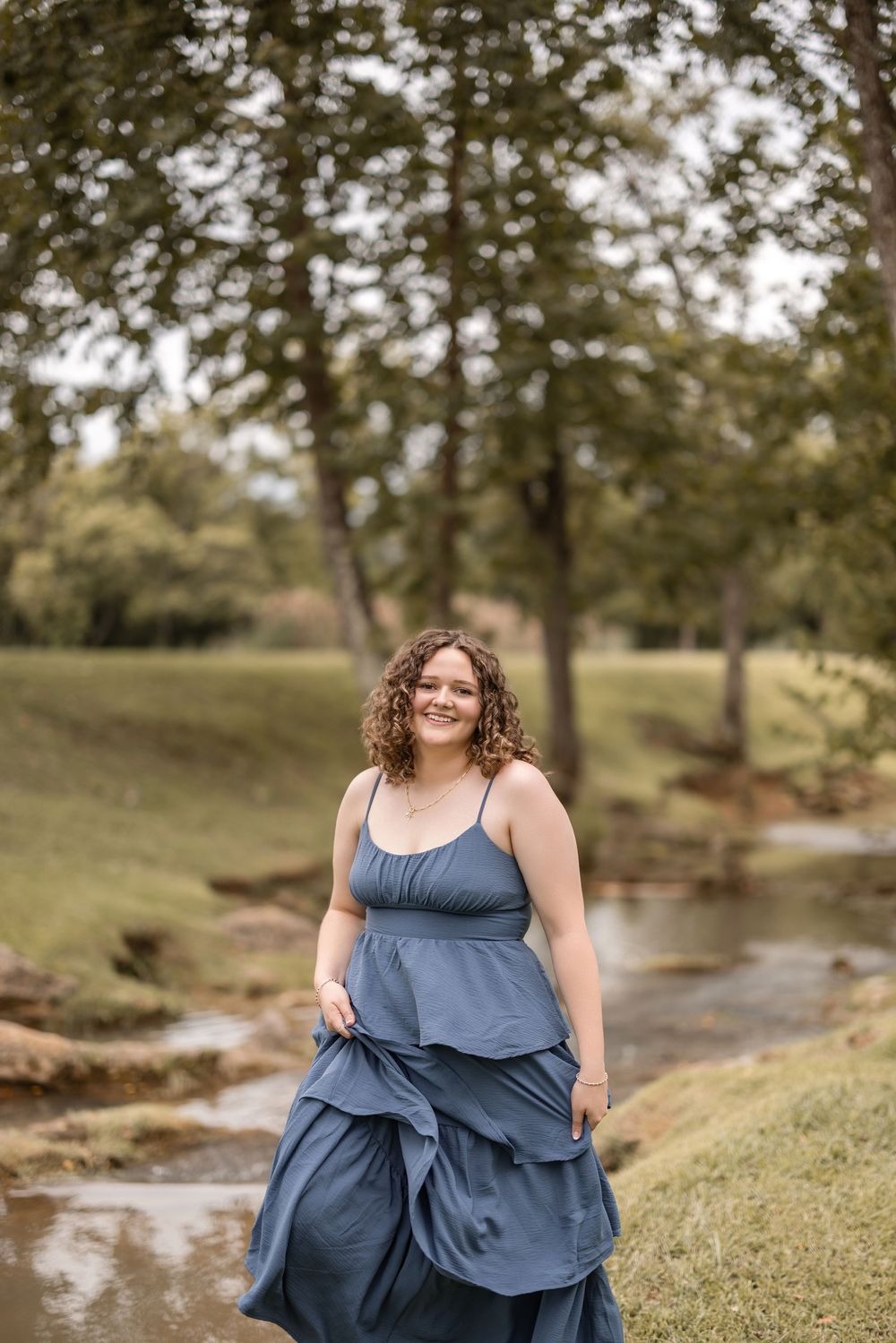 Someone in a flowing blue dress stands beside a peaceful stream in a wooded setting.