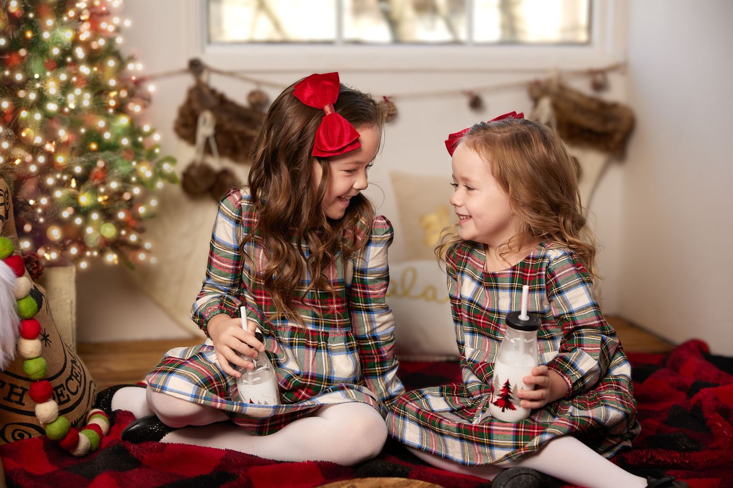 Two young sisters in matching plaid holiday dresses share a giggle by a Christmas tree with stockings in the background.