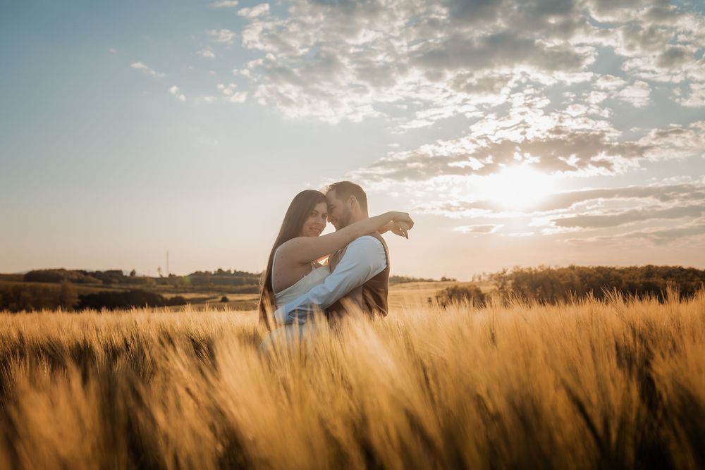 A romantic couple embraces in a golden wheat field at sunset with dramatic clouds in the sky.