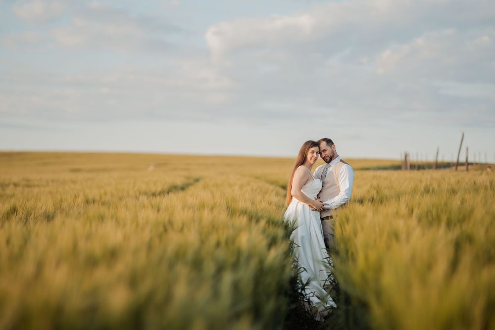 A romantic couple stands embracing in a golden wheat field at sunset with wispy clouds overhead.