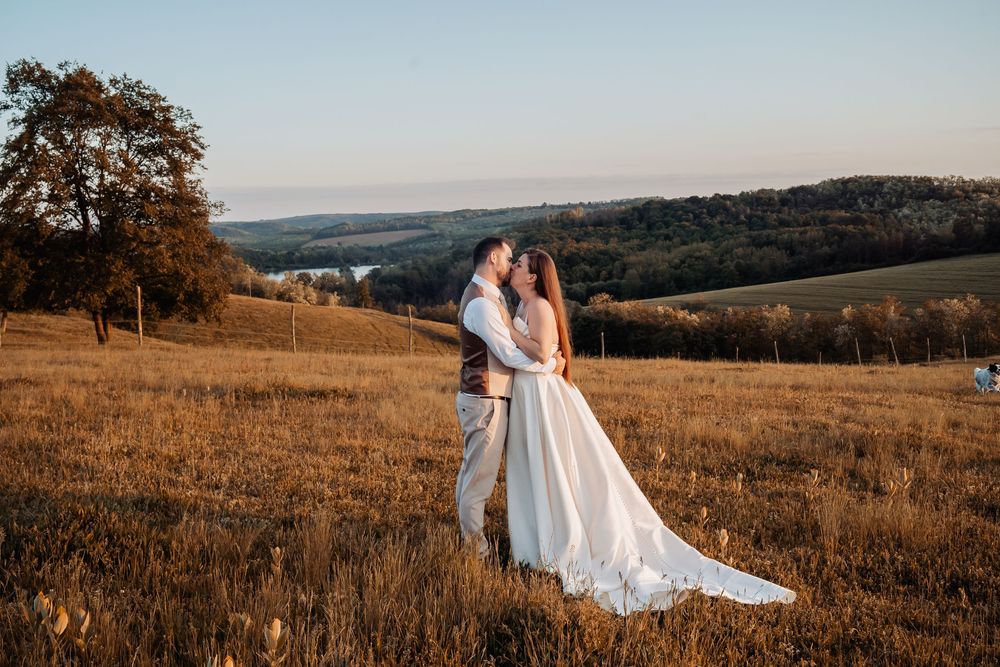 A couple embraces on a hilltop meadow during golden hour with rolling countryside landscape in the background.