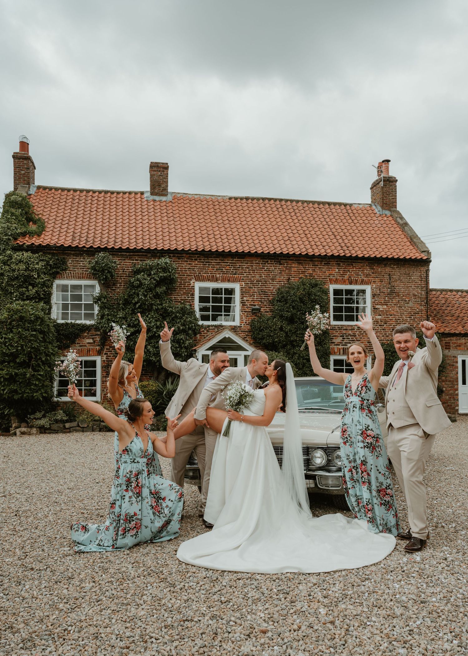 Wedding party celebrates with playful jumping poses outside a rustic brick farmhouse on a cloudy day.