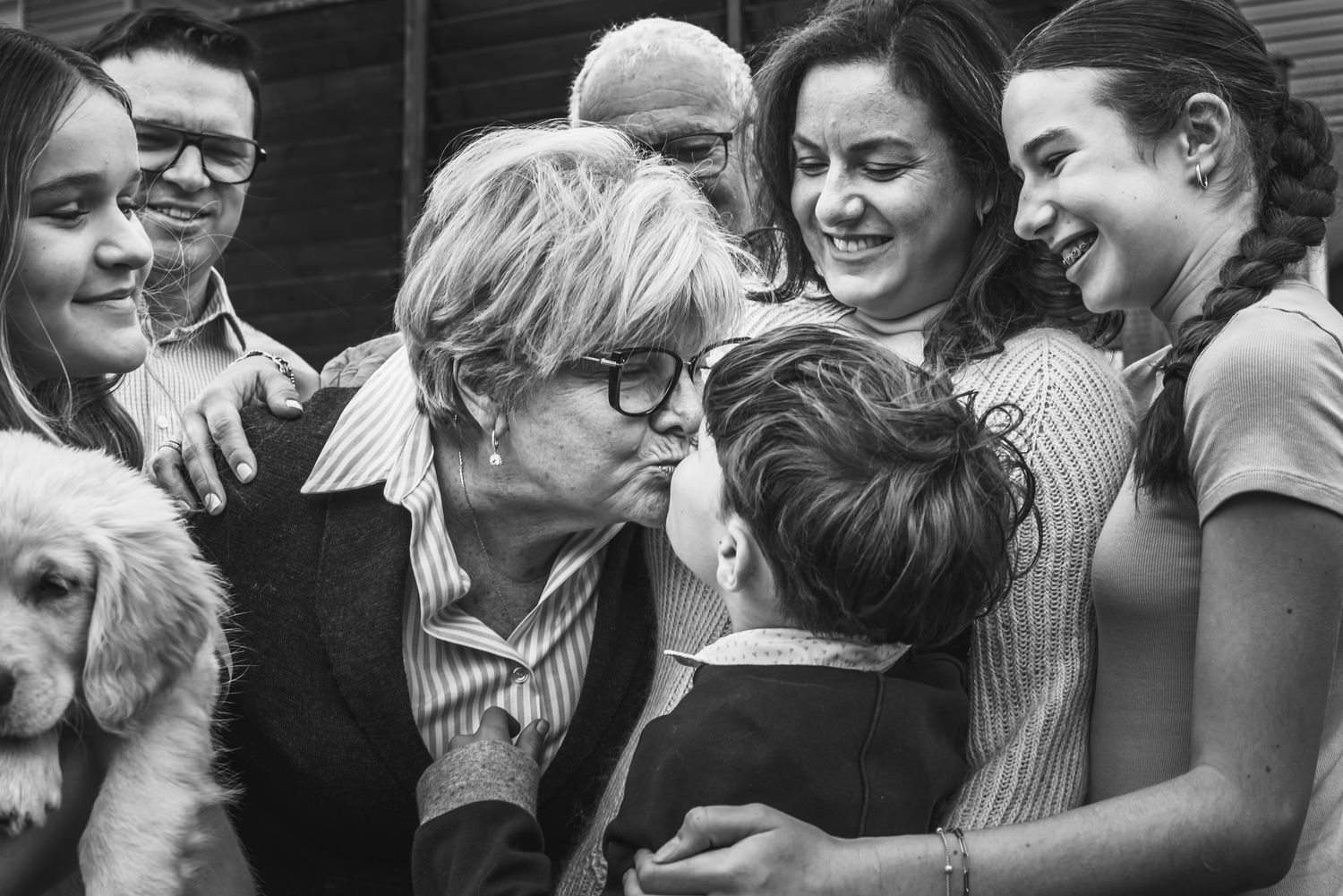 A group of people smile and laugh together as a grandma leans in to give her grandson a kiss.
