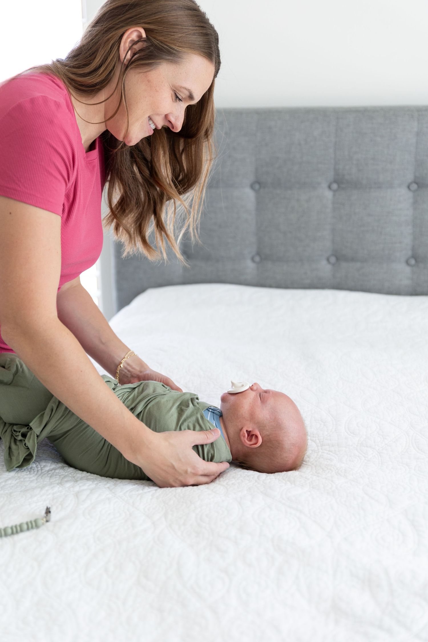 Mom in a bright pink shirt swaddles a crying baby on a white blanket bed