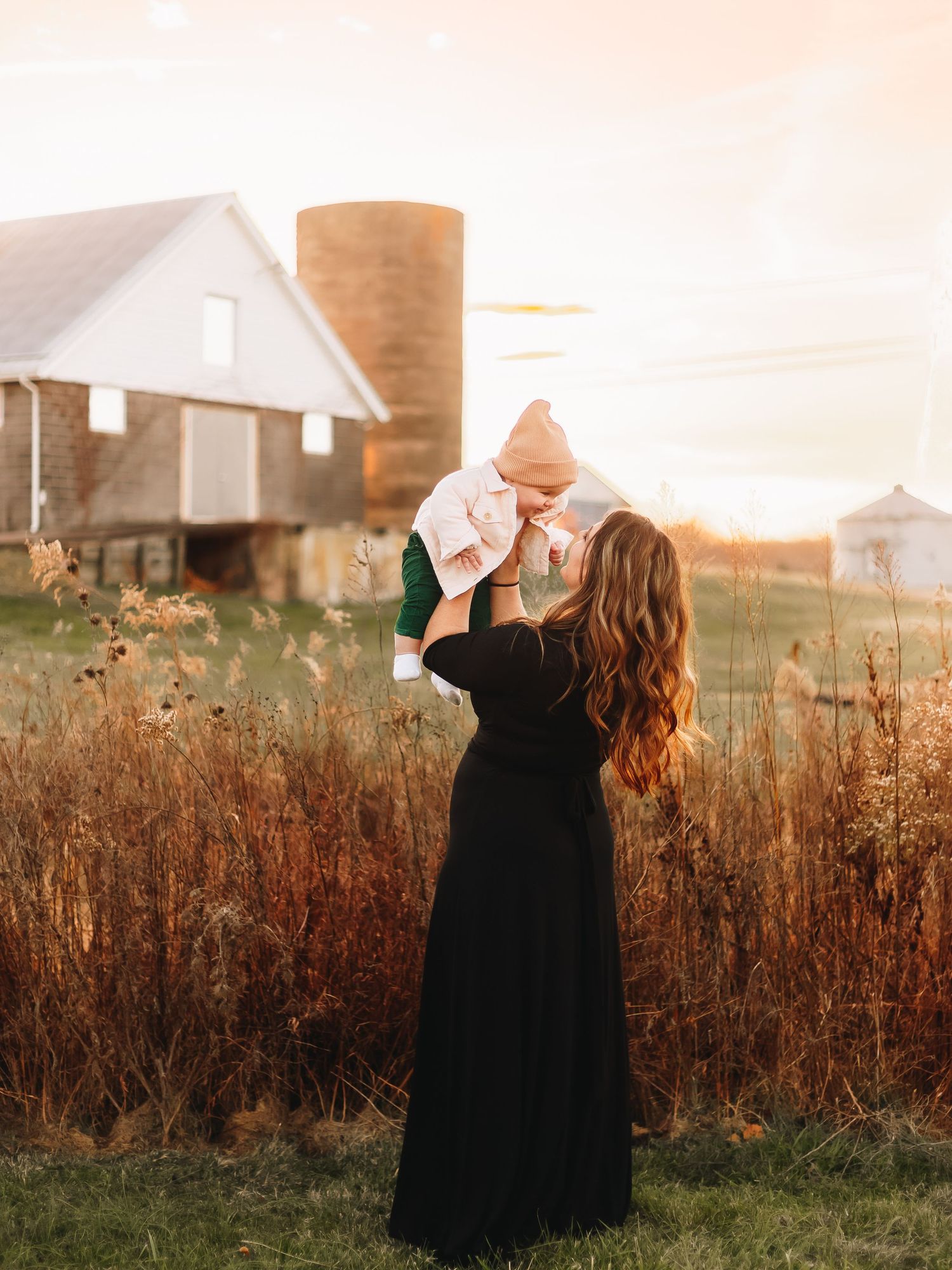Person in black dress spinning in a field at sunset with farm buildings in background.