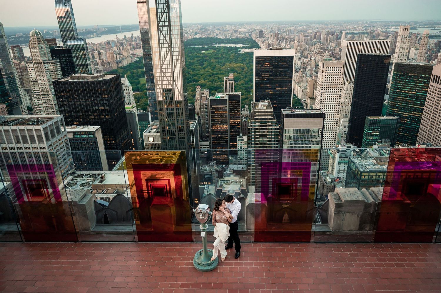 Couple embracing at observation deck overlooking Manhattan skyline through colorful glass panels.