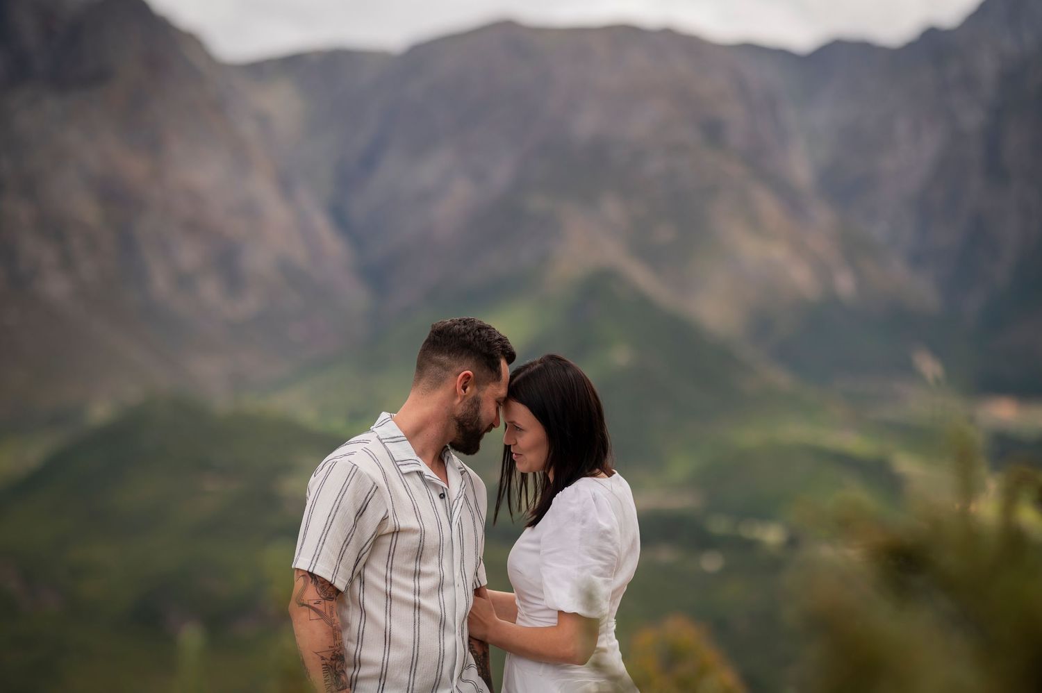 Two people embrace in front of scenic mountain landscape during outdoor photoshoot.