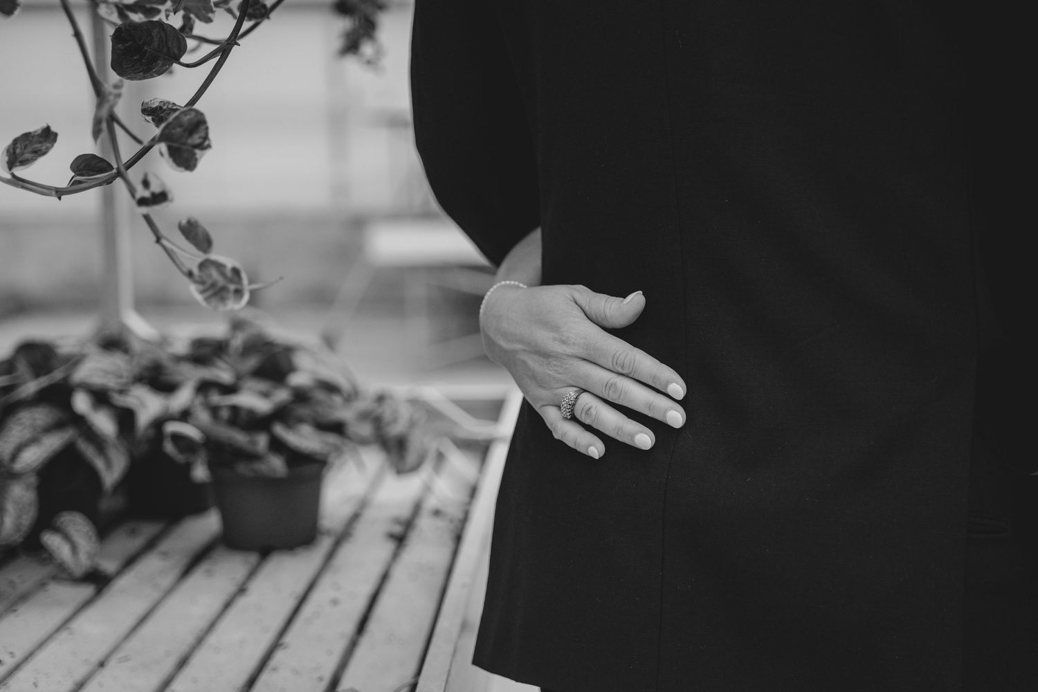 Black and white close-up of a hand against dark clothing with potted plants on a wooden surface in background.