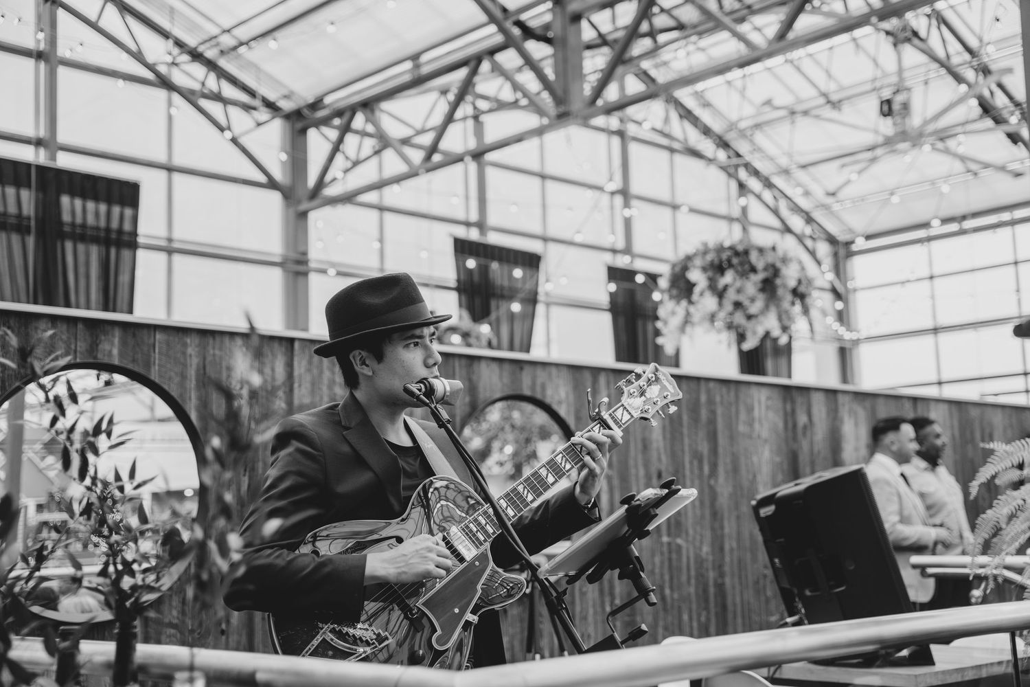 A performer plays an electric guitar while wearing a dark cap and jacket during an outdoor venue performance.