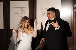 A bride and groom share a joyful dance moment together in formal attire against a wall with framed artwork.