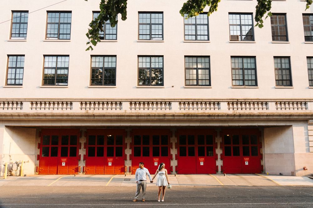 A couple holding hands stands in front of a beige building with bright red doors and rows of windows.