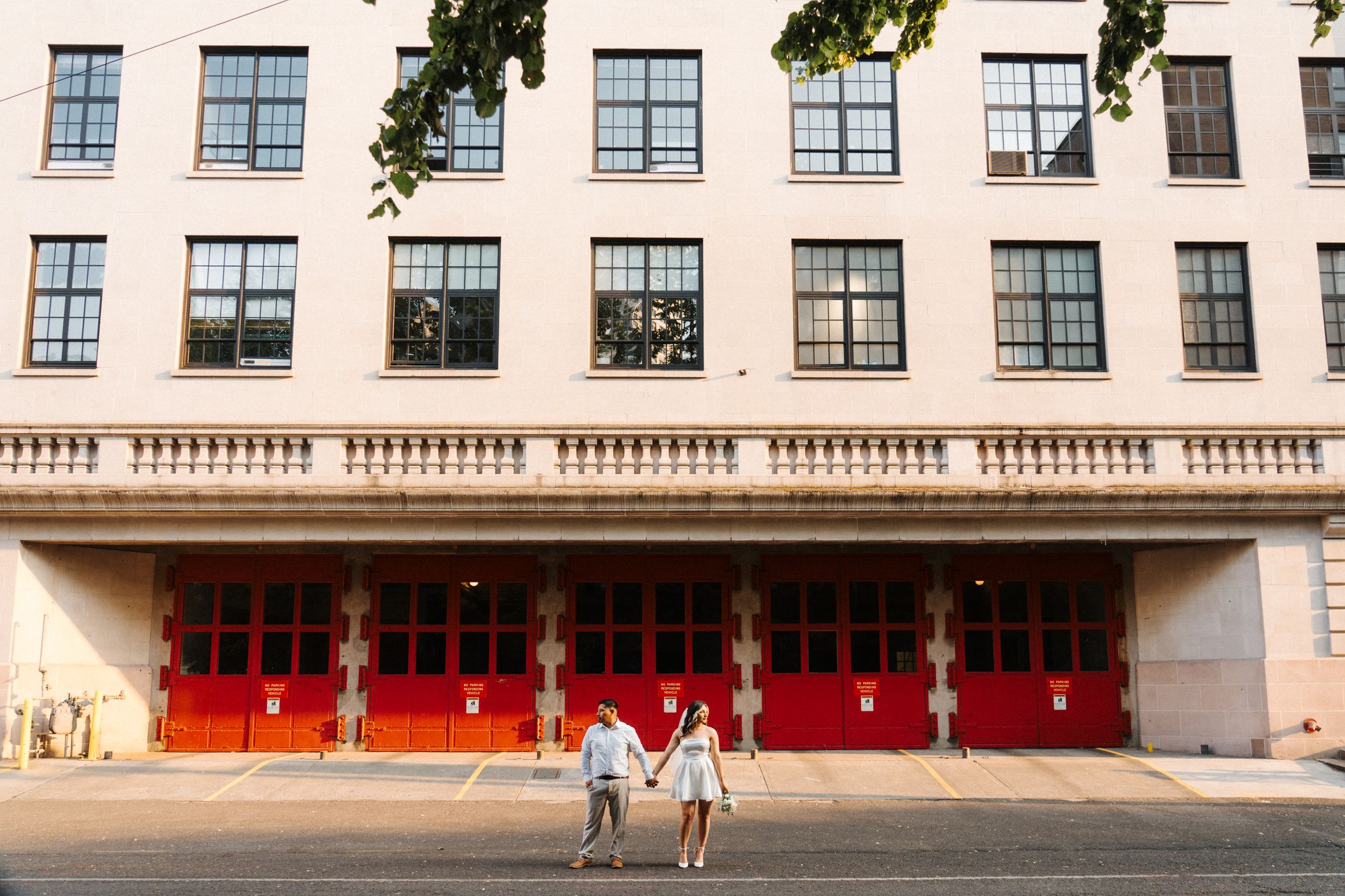 A couple holding hands stands in front of a beige building with bright red doors and rows of windows.