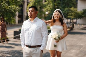 A couple in white wedding attire smile and pose together on an urban sidewalk on a sunny day.