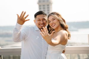 Happy couple in wedding attire showing off engagement ring on rooftop with cityscape in background.