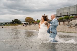 A wedding couple splashes joyfully in the water at a rocky beach under cloudy skies.