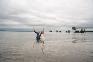 Couple celebrates in shallow ocean water with hands raised beneath overcast sky near coastal dock.