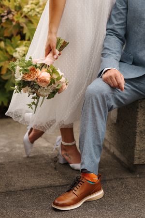 Detail shot of newlywed couple's feet and bridal bouquet showing white dress, gray suit, brown leather shoes and pink roses.