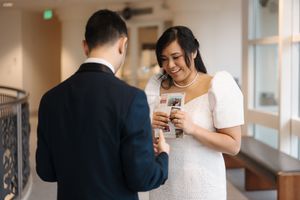 A couple getting ready for their wedding, checking their phones while dressed in a suit and white dress indoors.