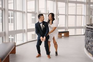 A couple laughs together in a bright indoor space, the bride wearing white and the groom in a dark suit.