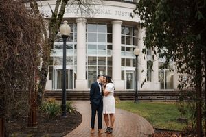 Couple standing together on brick pathway in front of elegant white columned building with large glass entryway.