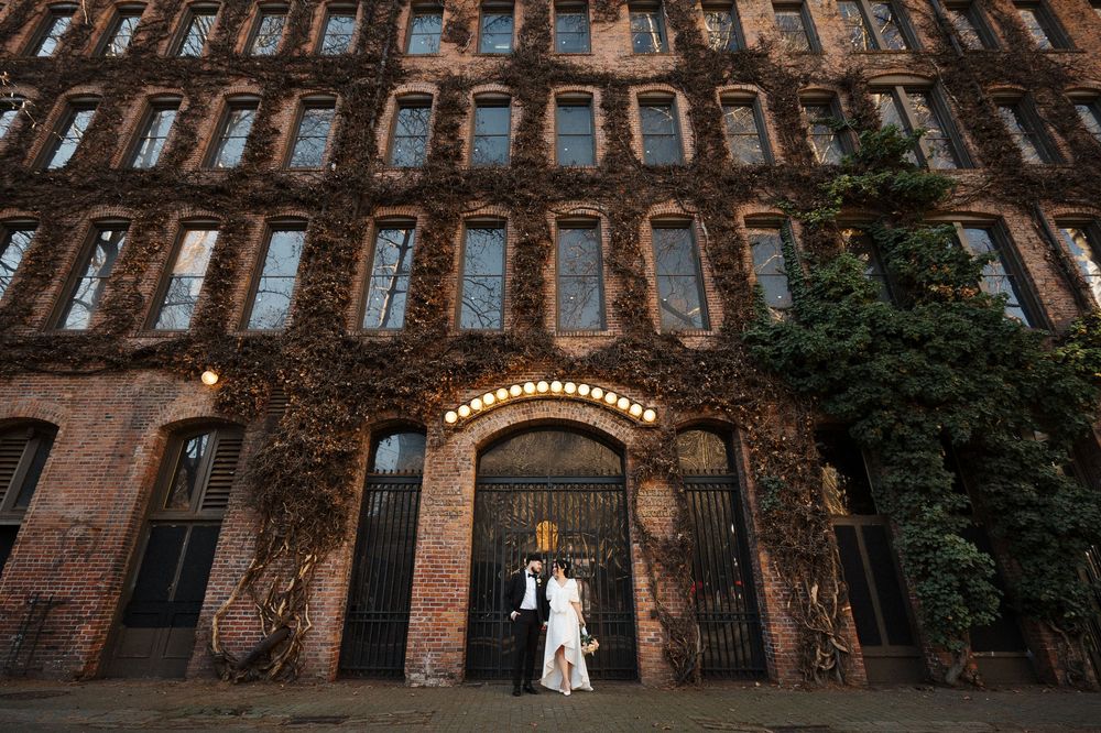 A rustic brick building exterior with arched windows and green foliage creates a charming wedding venue backdrop.