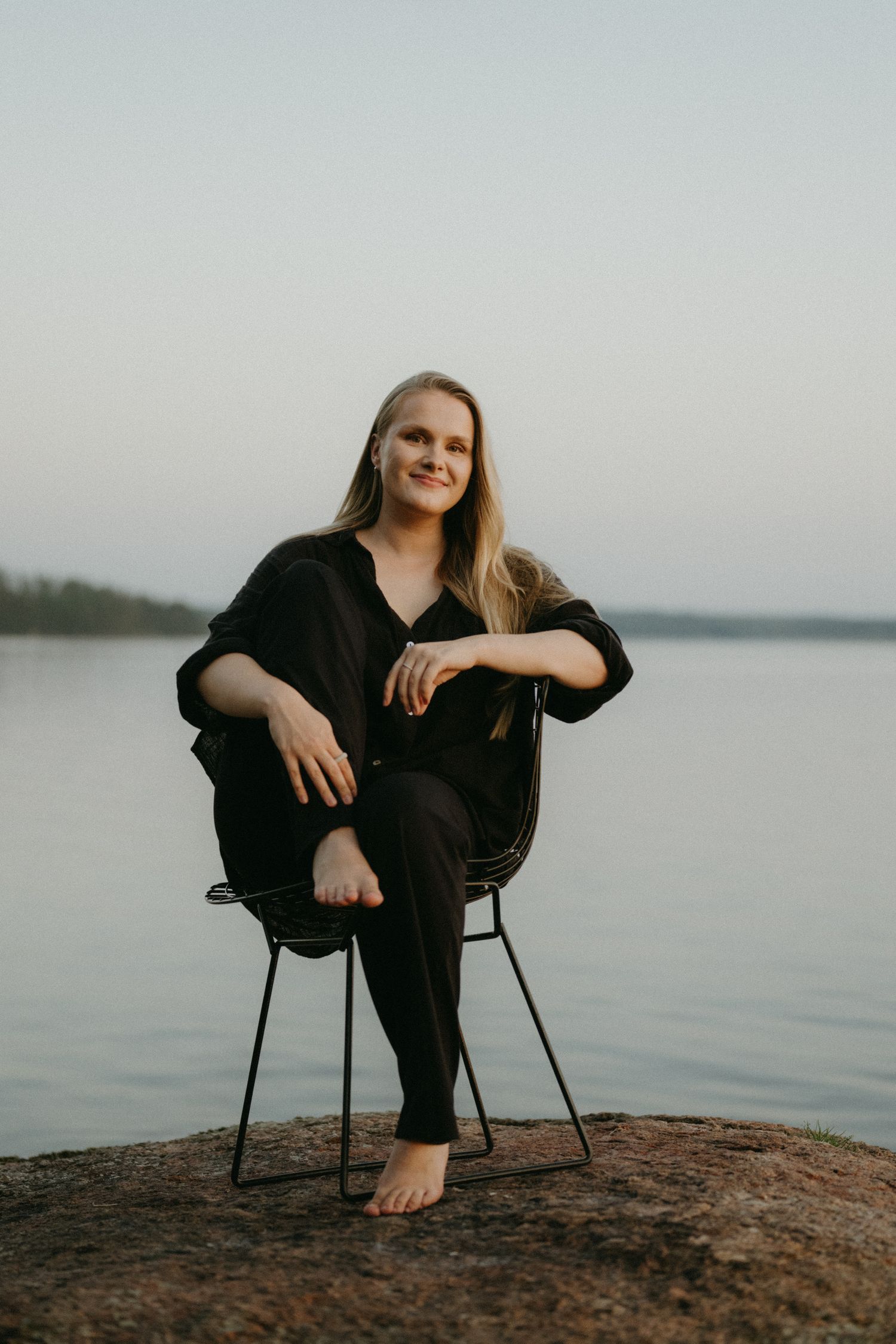 A figure in black clothing sits casually on a chair by a serene lakeside during an overcast day.