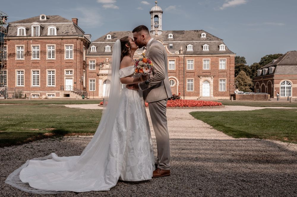 Couple in wedding attire embrace on pathway in front of grand red brick mansion with clock tower on summer day.
