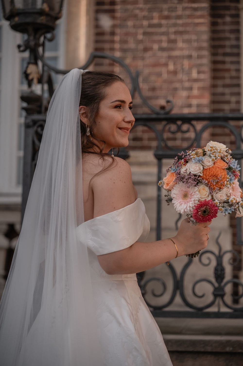 A bride in an off-shoulder white gown holds a vibrant bouquet while standing near ornate iron railings.