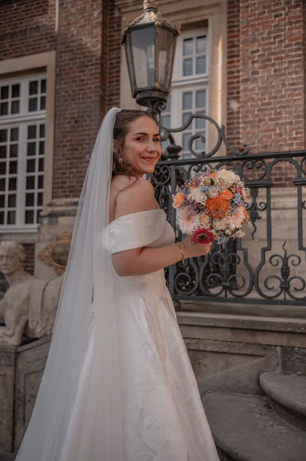 A bride in an elegant off-shoulder gown holds a colorful bouquet while standing by an ornate iron lamppost.