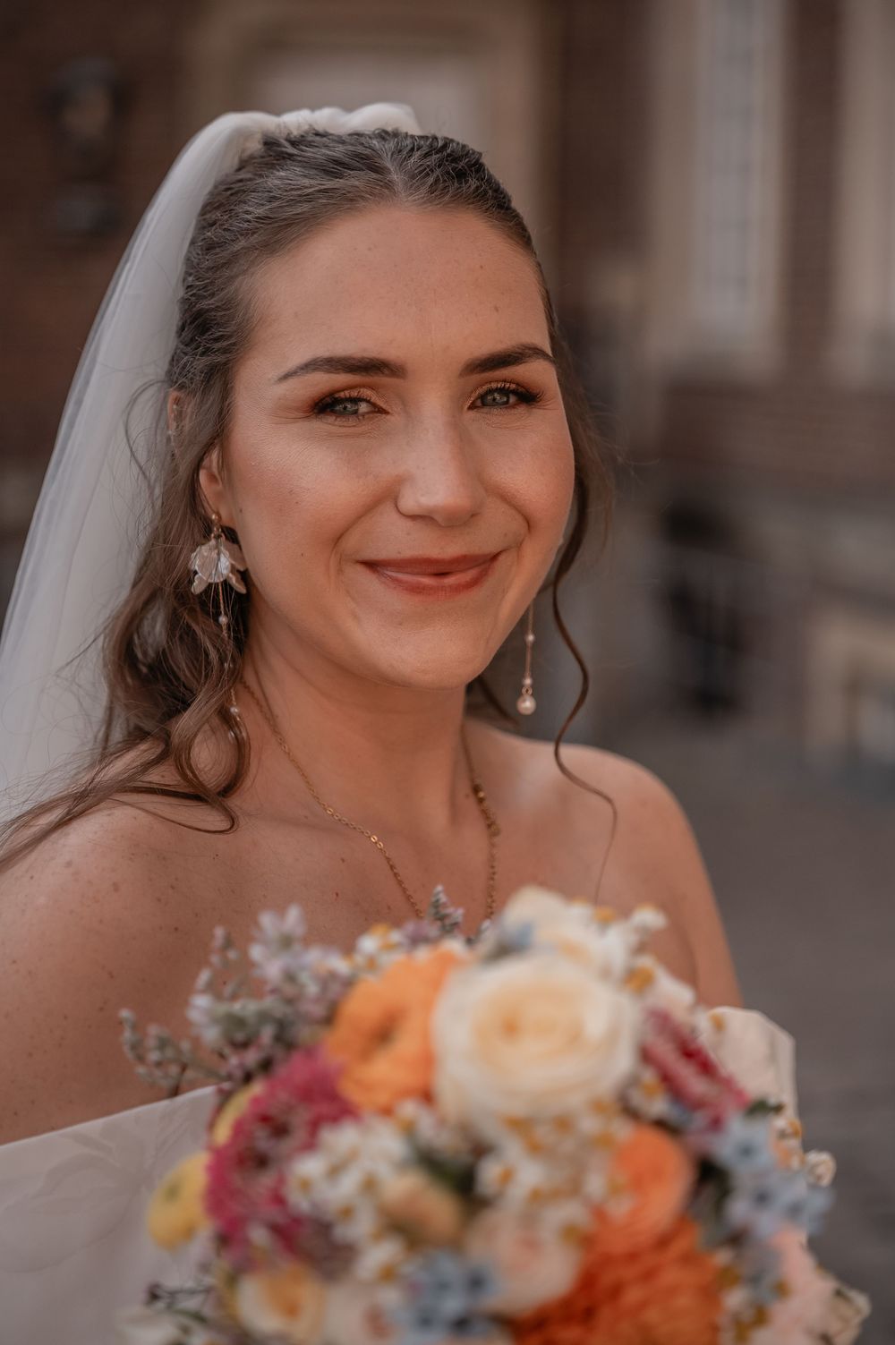 A radiant bride with dangling earrings holds a colorful bouquet featuring orange and pink flowers.