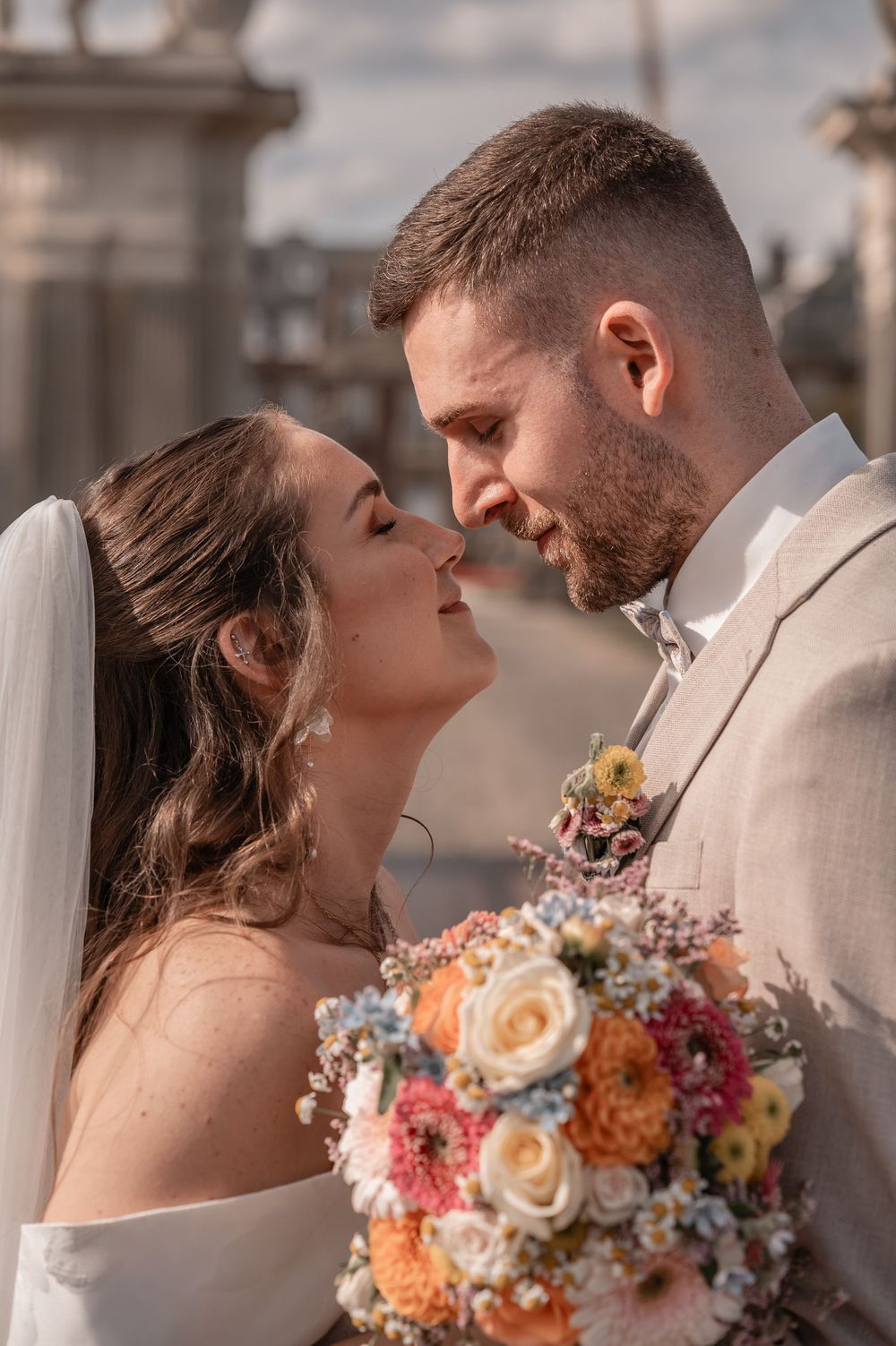 Wedding couple shares intimate moment outdoors, bride holding vibrant coral and peach bouquet.