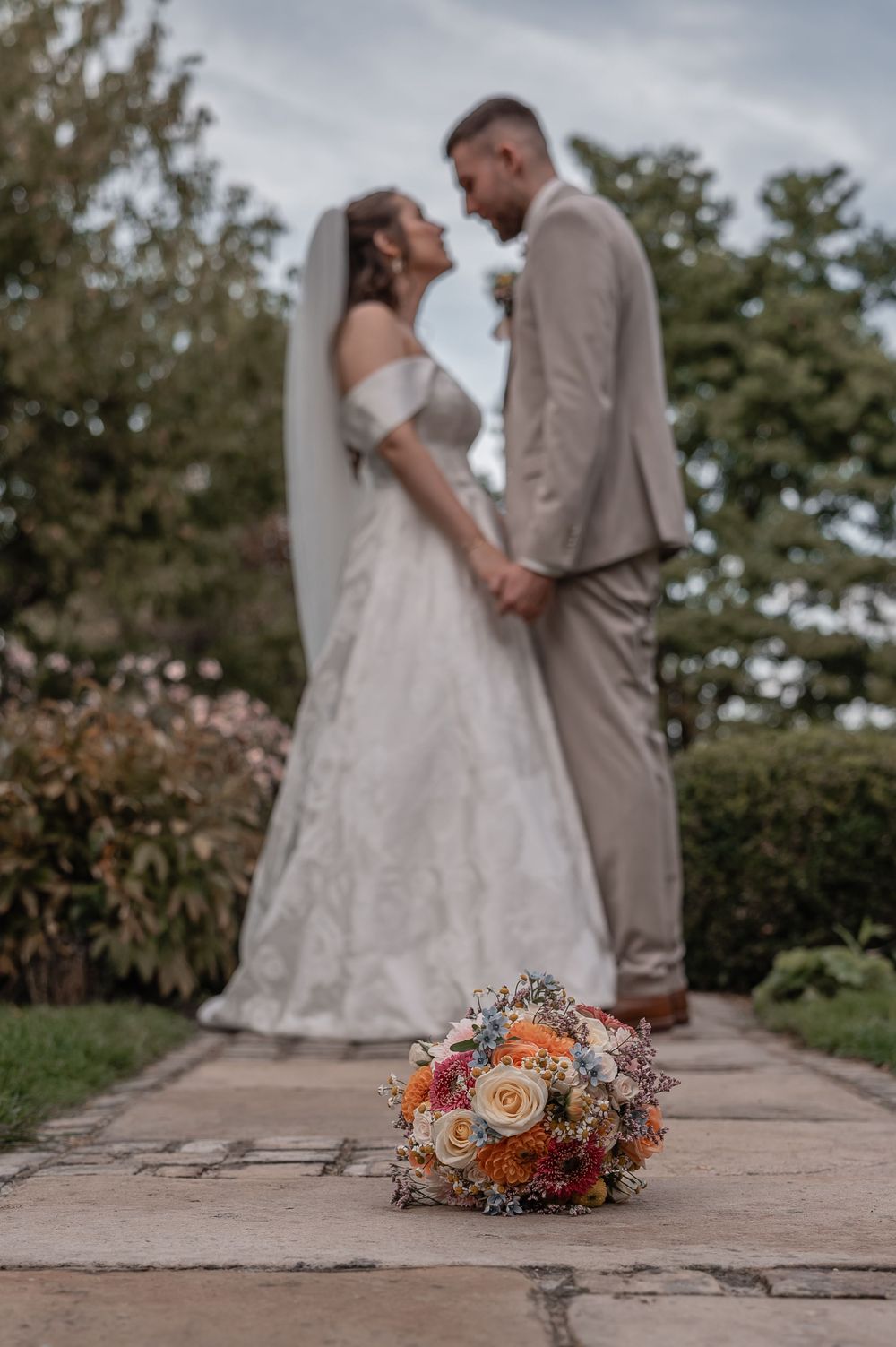 A beautiful wedding couple stands together on a stone path with vibrant fall floral bouquet in the foreground.